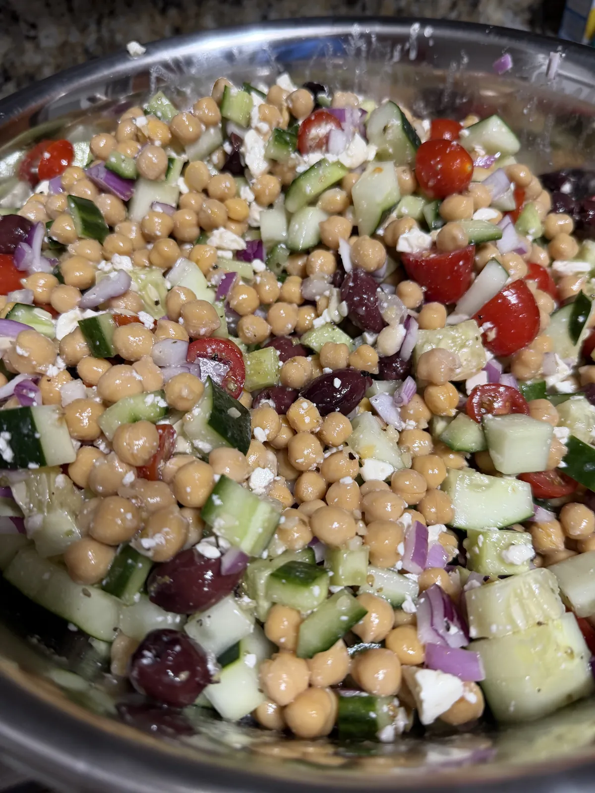 A steel bowl of tossed chickpeas, diced cucumber, halved cherry tomatoes, red onion, kalamata olives and crumbled feta