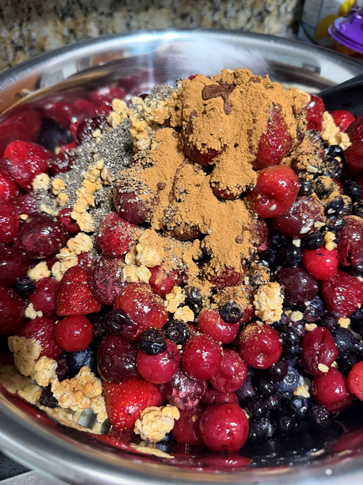 Frozen berries and cherries in a steel mixing bowl, topped with chia seeds, cinnamon and granola, before stirring